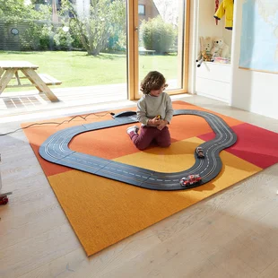 Teppich im Kinderzimmer - Ein Teppich in fröhlichen Farben liegt im Kinderzimmer. Ein Kind spielt darauf mit einer Rennbahn.